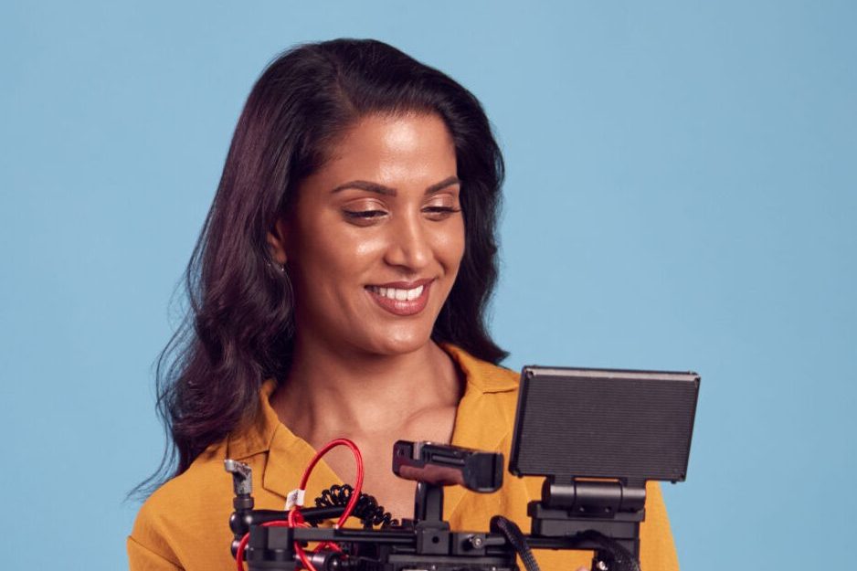 Mature Female Videographer With Camera Filming Video Against Blue Background On Shoot In Studio